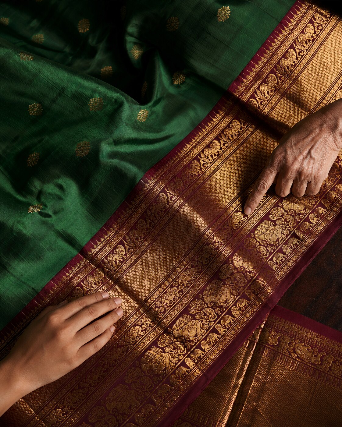 Older and younger hands appreciating the richly woven antique-gold zari border of an emerald and maroon Kanchipuram silk saree